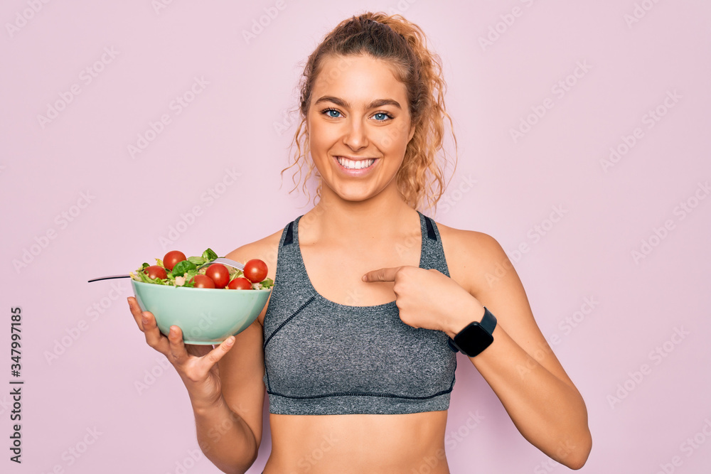 Young beautiful blonde sporty woman with blue eyes holding bowl with healthy salad with surprise face pointing finger to himself Young beautiful blonde sporty woman with blue eyes holding bowl with healthy salad with surprise face pointing finger to himself