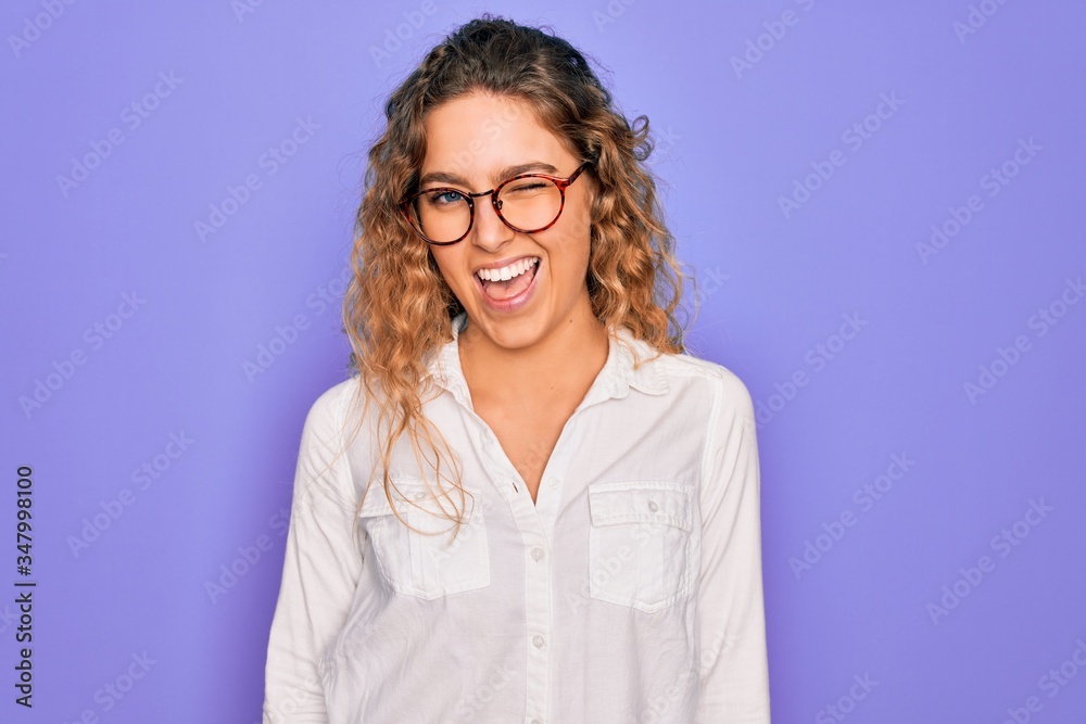 Young beautiful woman with blue eyes wearing casual shirt and glasses over purple background winking looking at the camera with sexy expression, cheerful and happy face.