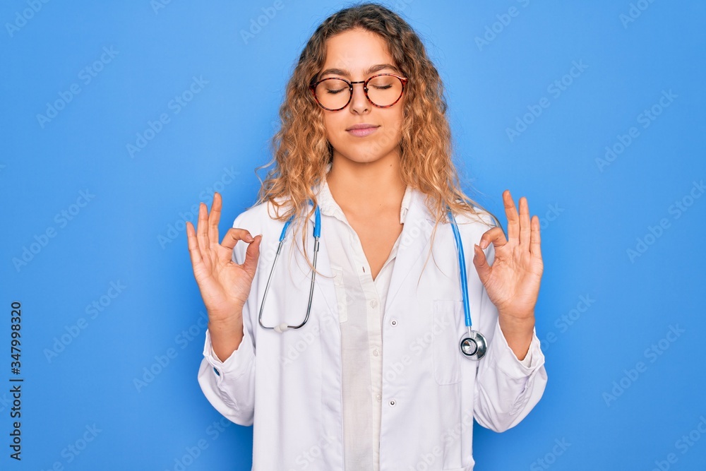 Young beautiful blonde doctor woman with blue eyes wearing coat and stethoscope relax and smiling with eyes closed doing meditation gesture with fingers. Yoga concept. Young beautiful blonde doctor woman with blue eyes wearing coat and stethoscope relax and smiling with eyes closed doing meditation gesture with fingers. Yoga concept.