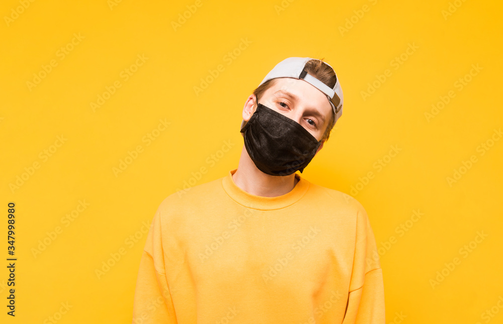 The guy in a cap and a black medical mask stands on a yellow background and poses for the camera with a serious face. Portrait of a young man in a protective gauze mask. Isolated. Coronavirus pandemic