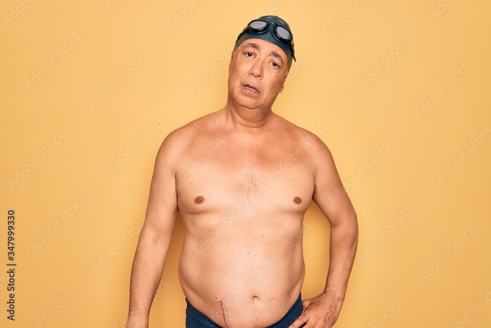 Middle age senior grey-haired swimmer man wearing swimsuit, cap and goggles In shock face, looking skeptical and sarcastic, surprised with open mouth