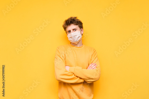 Portrait of an emotional guy in a medical mask and a yellow sweatshirt stands on a yellow background and looks at the camera with a displeased face. Coronavirus pandemic. Quarantine. covid-19.