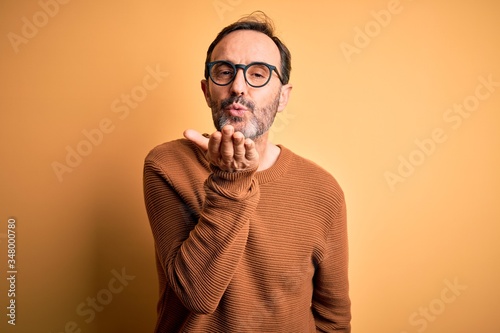 Middle age hoary man wearing brown sweater and glasses over isolated yellow background looking at the camera blowing a kiss with hand on air being lovely and sexy. Love expression.