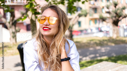 Blonde Young girl in yellow sunglasses, red lipstick taking sun baths and vitamin D from sunlight. Green park backdrop. Summer positive vibes concept, modern lifestyle, sunshine weather