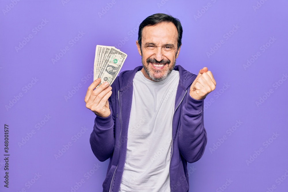 Middle age handsome man holding bunch of dollars banknotes over isolated purple background screaming proud, celebrating victory and success very excited with raised arm Middle age handsome man holding bunch of dollars banknotes over isolated purple background screaming proud, celebrating victory and success very excited with raised arm