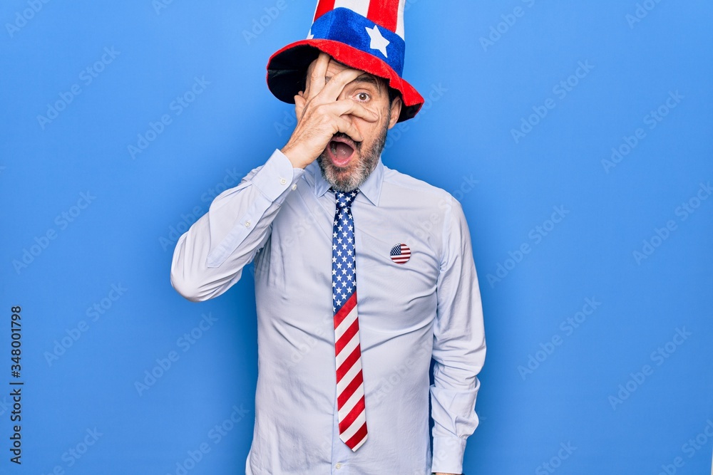 Middle age handsome patriotic man wearing united states hat and tie over blue background peeking in shock covering face and eyes with hand, looking through fingers afraid Middle age handsome patriotic man wearing united states hat and tie over blue background peeking in shock covering face and eyes with hand, looking through fingers afraid