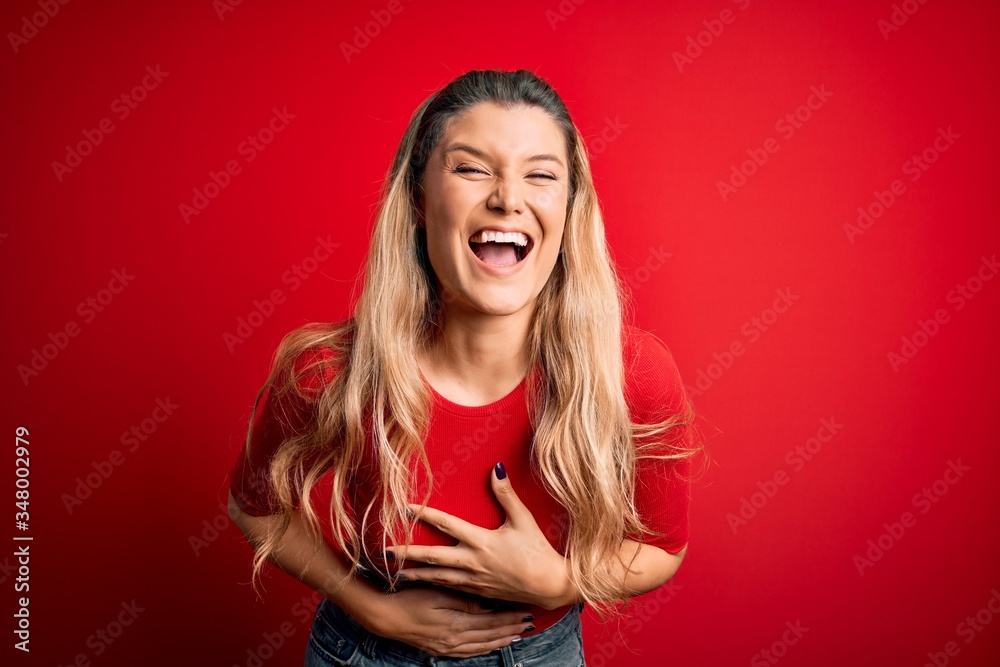 Young beautiful blonde woman wearing casual t-shirt standing over isolated red background smiling and laughing hard out loud because funny crazy joke with hands on body. Young beautiful blonde woman wearing casual t-shirt standing over isolated red background smiling and laughing hard out loud because funny crazy joke with hands on body.