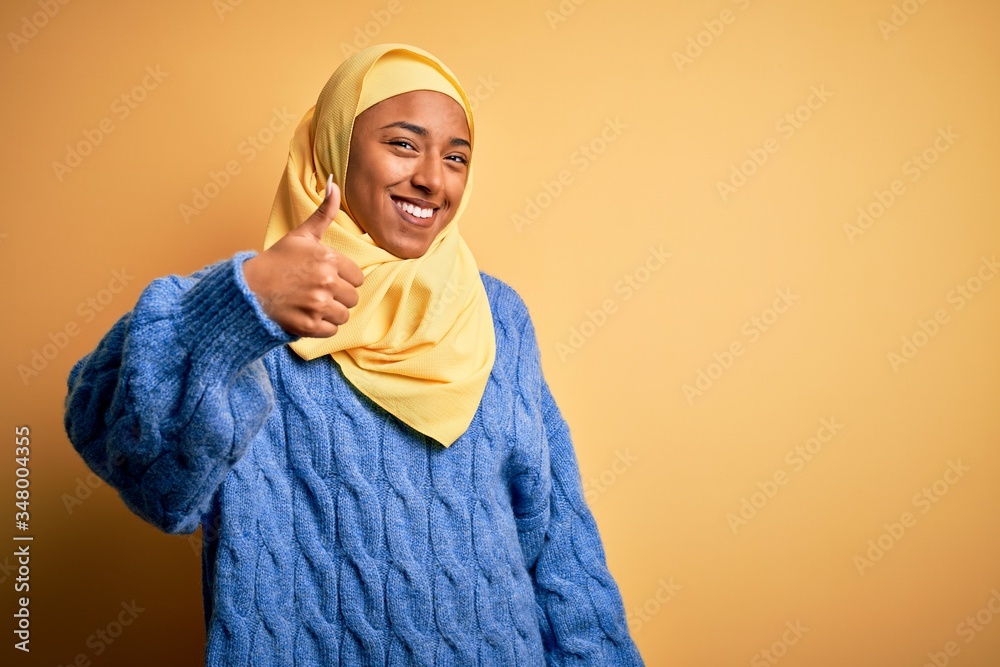 Young African American afro woman wearing muslim hijab over isolated yellow background doing happy thumbs up gesture with hand. Approving expression looking at the camera showing success. Young African American afro woman wearing muslim hijab over isolated yellow background doing happy thumbs up gesture with hand. Approving expression looking at the camera showing success.