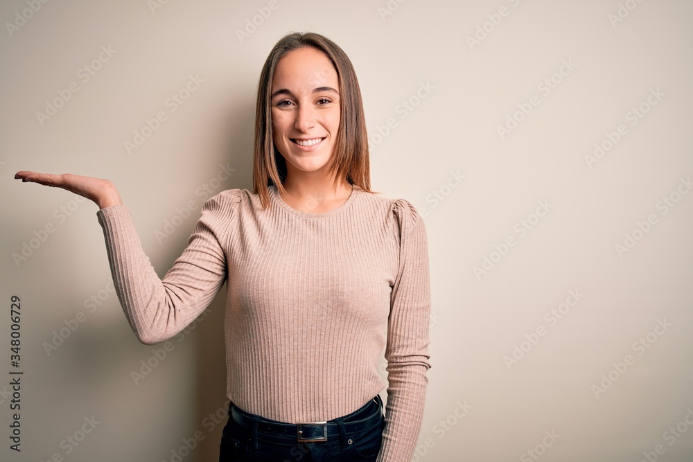 Young beautiful woman wearing casual sweater standing over isolated white background smiling cheerful presenting and pointing with palm of hand looking at the camera. Young beautiful woman wearing casual sweater standing over isolated white background smiling cheerful presenting and pointing with palm of hand looking at the camera.