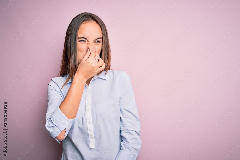 Young beautiful businesswoman wearing elegant shirt standing over isolated pink background smelling something stinky and disgusting, intolerable smell, holding breath with fingers on nose. Bad smell Young beautiful businesswoman wearing elegant shirt standing over isolated pink background smelling something stinky and disgusting, intolerable smell, holding breath with fingers on nose. Bad smell