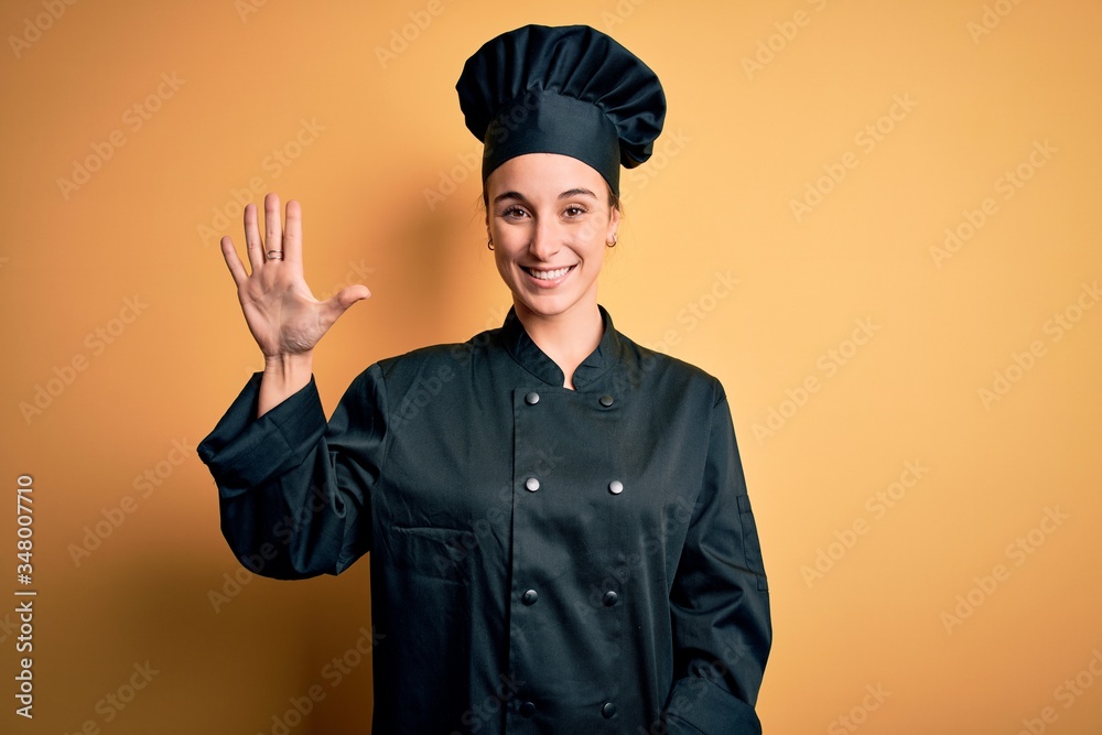 Young beautiful chef woman wearing cooker uniform and hat standing over yellow background showing and pointing up with fingers number five while smiling confident and happy.