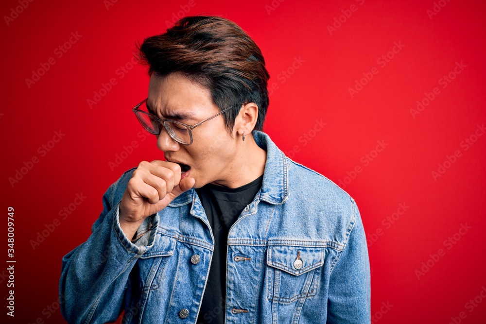 Young handsome chinese man wearing denim jacket and glasses over red background feeling unwell and coughing as symptom for cold or bronchitis. Health care concept.