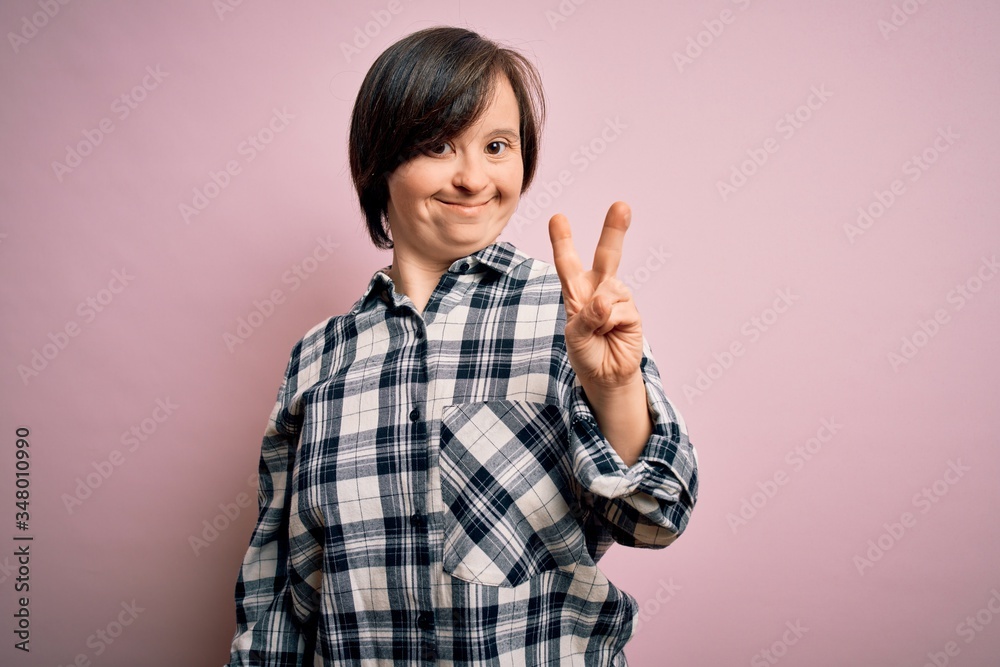 Young down syndrome woman wearing casual shirt over pink background smiling with happy face winking at the camera doing victory sign. Number two. Young down syndrome woman wearing casual shirt over pink background smiling with happy face winking at the camera doing victory sign. Number two.