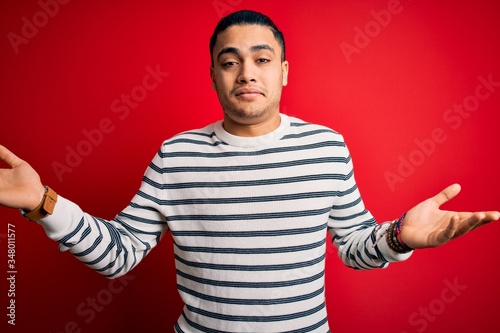 Young brazilian man wearing casual striped t-shirt standing over isolated red background clueless and confused expression with arms and hands raised. Doubt concept.