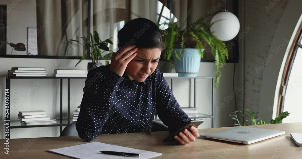 Confused unhappy indian businesswoman feels frustrated about broken phone problem in office. Annoyed female worker reading bad news in message, perplexed by mobile spam sms notification at workplace.