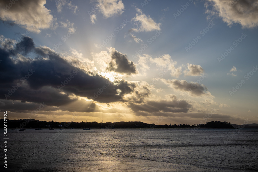 sunset over the ocean with the sun behind clouds and sun beams around the clouds and boats in the water