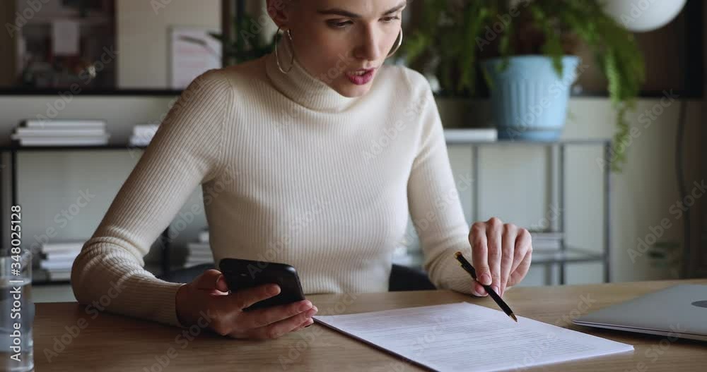 Focused young business woman student worker using smartphone checking paper sitting at home office desk. Female interpreter translator working studying translating document using mobile elearning app.