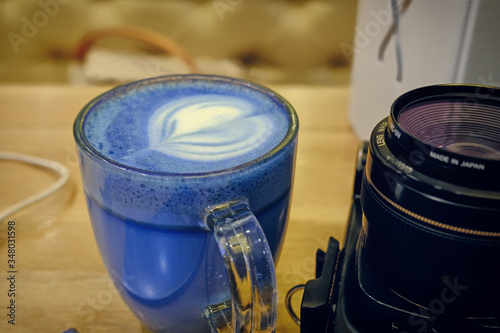 Trendy drink: Blue latte. Top view of hot classic blue color butterfly pea latte tea or blue spirulina latte and photo camera nearby on wood textured background.