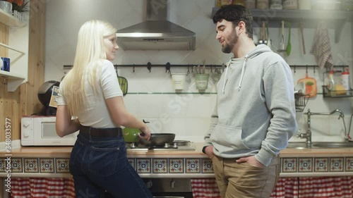 Young wife cooks something on pan and her husband talks to her photo