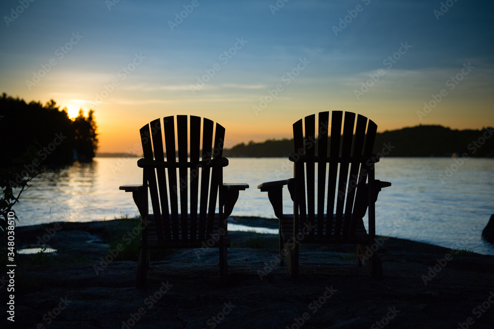 Silhouette of two Adirondack chairs sit on a rock formation facing the calm waters of a lake during a sunset in Muskoka, Ontario Canada. 