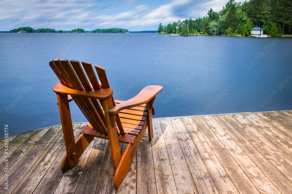 Adirondack chair sitting on a wooden dock facing a blue calm lake. Across the water is a white cottage nestled between green trees. 