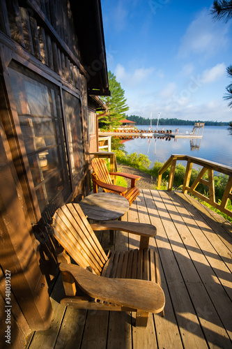 Chairs sit on a cottage wooden porch during a summer day in Canada.  A wooden dock, populated with lounge chairs is visible in the background. Sailing boats and people activity are also in the frame.