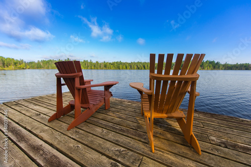 Two Muskoka chairs sitting on a wood dock facing a lake.