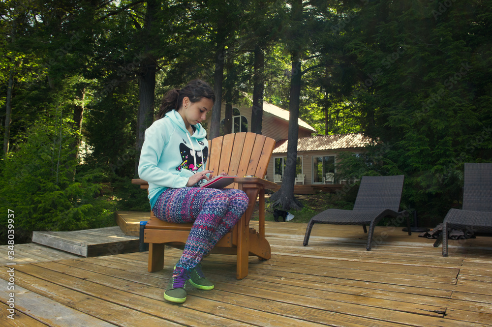 Fototapeta premium Teenager girl sit on a Muskoka chair reading on a tablet. The girl is wearing colourful clothes. A cottage nestled between trees is visible in the background.