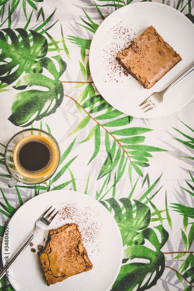 Top view of a cup of coffee and two dessert plates with brownie cake over table-cloth with green leaves pattern.