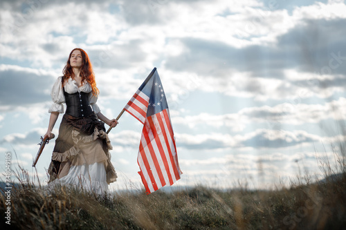 Girl in historical dress of 18th century with flag of United States. July 4 is US Independence Day. Woman of patriot freedom fighter in outdoor on background cloudy sky photo