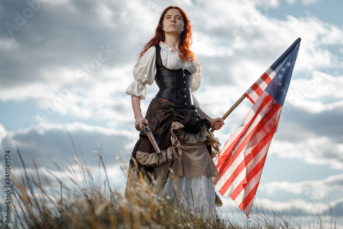 Girl in historical dress of 18th century with flag of United States. July 4 is US Independence Day. Woman of patriot freedom fighter in outdoor on background cloudy sky photo