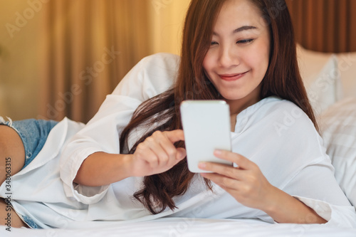 Closeup image of a beautiful asian woman using and looking at mobile phone while lying down on a white bed