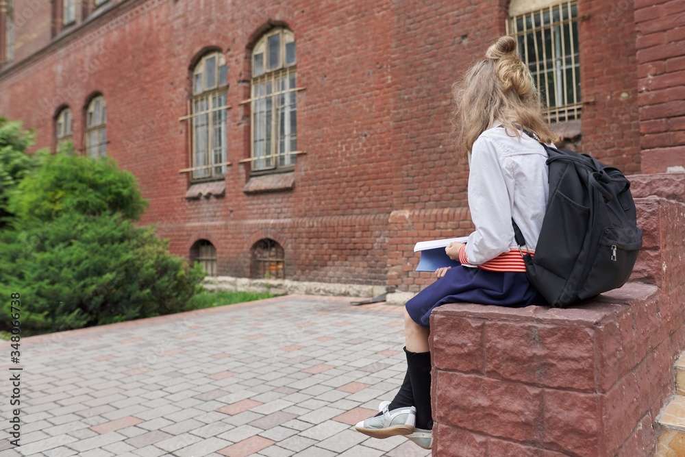 Girl child with backpack sitting in school yard reading notebook, copy space