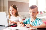 Boy and girl, brother and sister study at home. A girl is watching a video lesson or an online conference, the boy is doing exercises in a notebook from a textbook. Boy looking at the camera