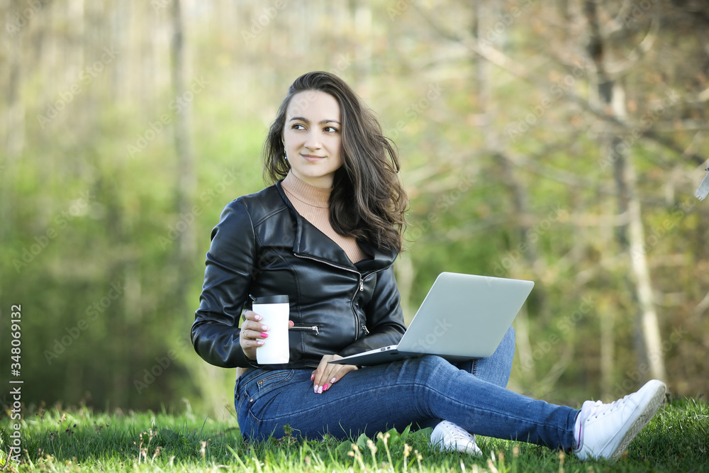 Young woman in a black jacked with coffee cup and laptop working outside in a park. Remote work. Digital work. Distance learning