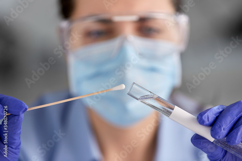 medicine, health and virus concept - close up of young female doctor or scientist in face protective medical mask, goggles and gloves holding beaker with coronavirus test and cotton swab