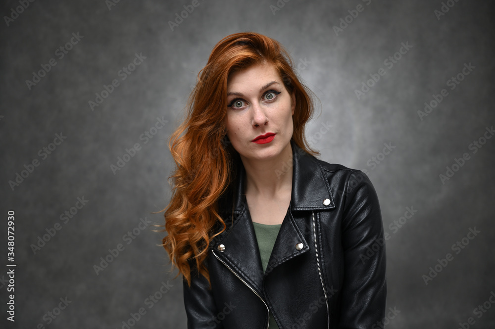 Studio photo of a young caucasian woman with long red hair in a black jacket. Pretty model actress posing with different emotions on a gray background in the studio. Studio photo of a young caucasian woman with long red hair in a black jacket. Pretty model actress posing with different emotions on a gray background in the studio.