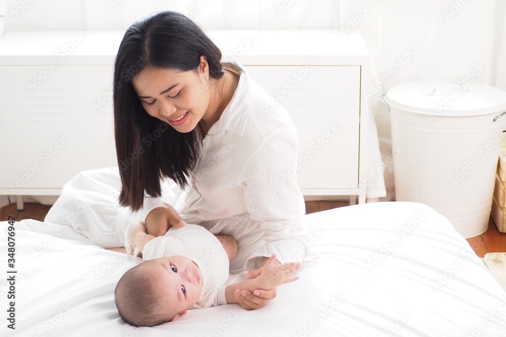 Beautiful Asian mother looking at and playing with her cute baby lying on bed in the bedroom in white and warm tone. Happy motherhood, unconditional love, family lifestyle concept Beautiful Asian mother looking at and playing with her cute baby lying on bed in the bedroom in white and warm tone. Happy motherhood, unconditional love, family lifestyle concept