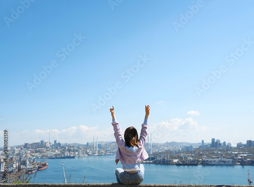  brunette girl in a purple denim jacket and blue jeans raises her hands up and looks at a large modern sea city with a port and cable-stayed bridge. rear view, freedom of movement, end of quarantine