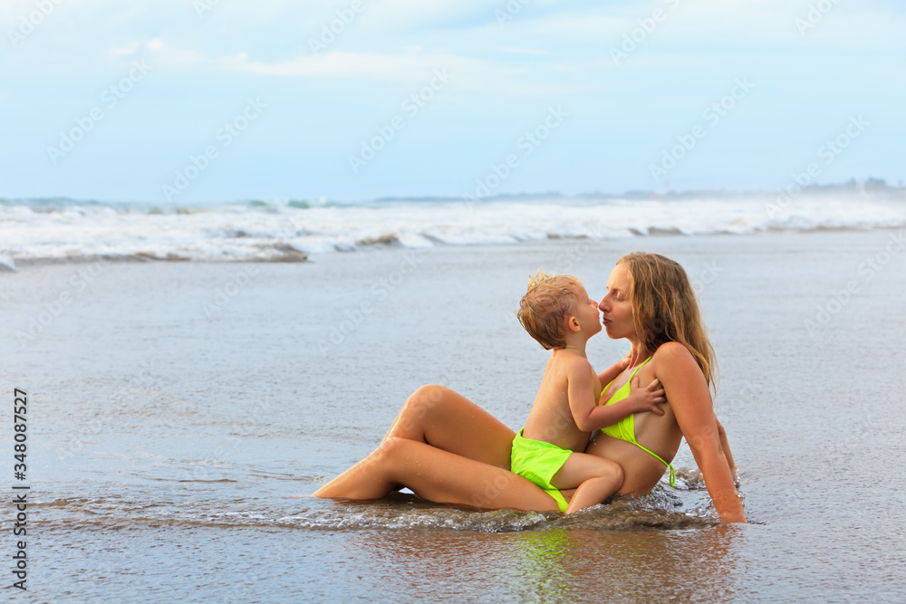 Fototapeta premium Happy family have fun on tropical sea beach resort. Funny baby boy cuddle and kiss young beautiful mother. Active kids lifestyle, people swimming activity on summer holiday with children.