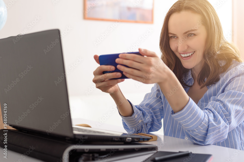 smiling cheerful woman in blue shirt with smartphone in hands, playing online games on phone, freelancer, video calls concept, remote work