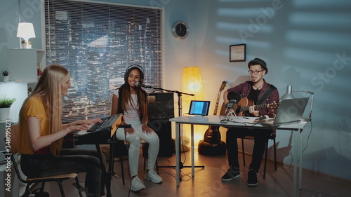 Multiracial amateur band is rehearsing by singing playing keyboard and guitar in home studio with cozy interior in the evening