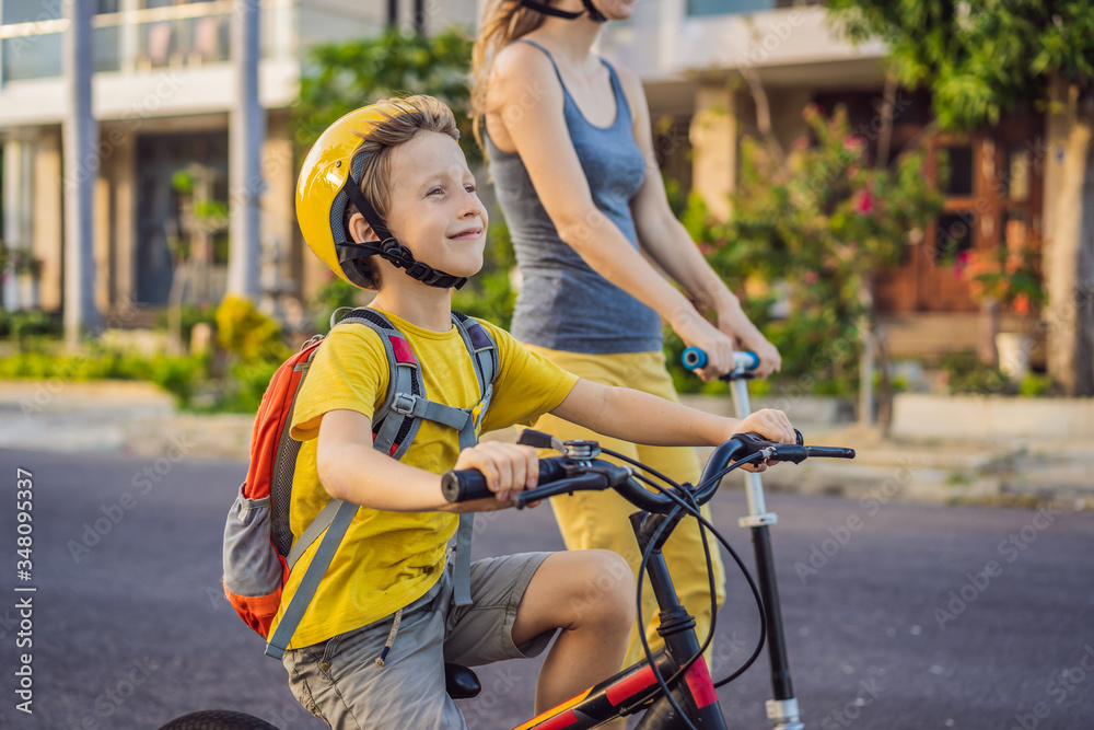 Obraz premium Active school kid boy and his mom in safety helmet riding a bike with backpack on sunny day. Happy child biking on way to school. Safe way for kids outdoors to school