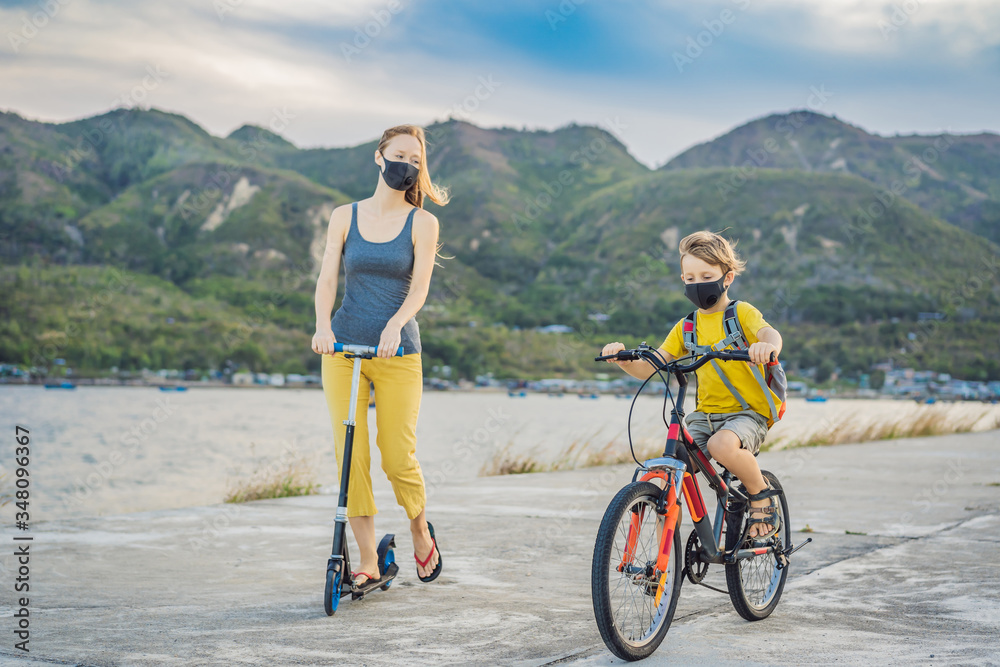 Fototapeta premium Active school kid boy and his mom in medical mask riding a bike with backpack on sunny day. Happy child biking on way to school. You need to go to school in a mask because of the coronavirus epidemic