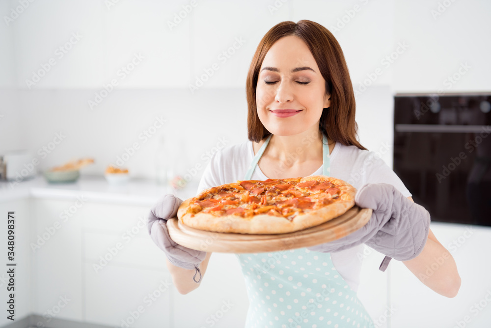 Photo of beautiful cheerful housewife enjoy hobby cooking family recipe dinner pizza smelling hot ready meal from oven wear t-shirt apron gloves standing modern kitchen indoors