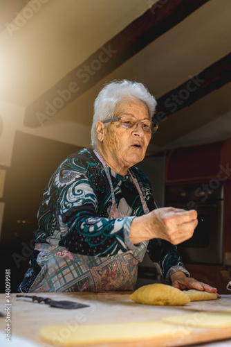 Caucasian old woman talking while cooking in her kitchen traditional recipes of Italian cuisine. Fresh handmade pasta with eggs and flour, rolling dough on wooden cutting board.