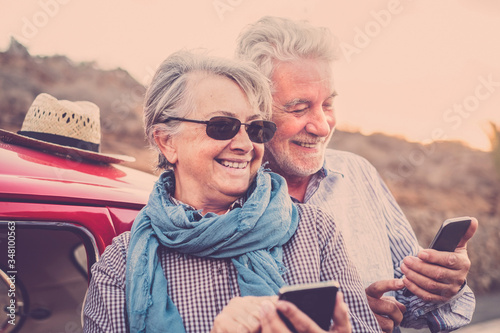 Senior happy cheerful old caucasian couple use together modern online technology smart phone to share and send contents  on the web - retired people outdoor enjoy life - red car in background photo