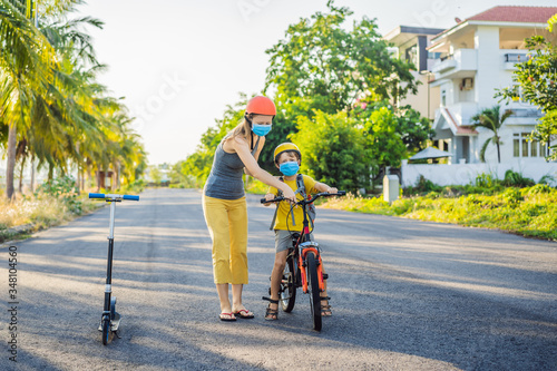 Active school kid boy and his mom in medical mask and safety helmet riding a bike with backpack on sunny day. Happy child biking on way to school. You need to go to school in a mask because of the