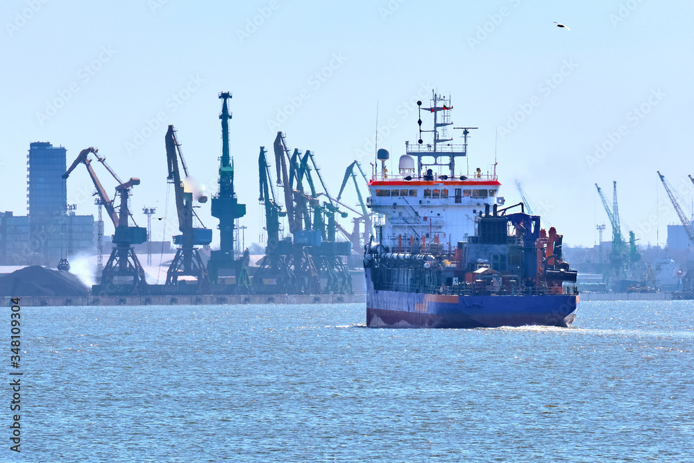 View of commercial cargo port with container bridge cranes and a ship entering port on a sunny day.