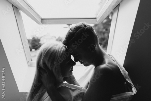 Young man and woman in sheets hug and kiss near the window. Black and white photo of a naked man and woman by the window. © Oksana
